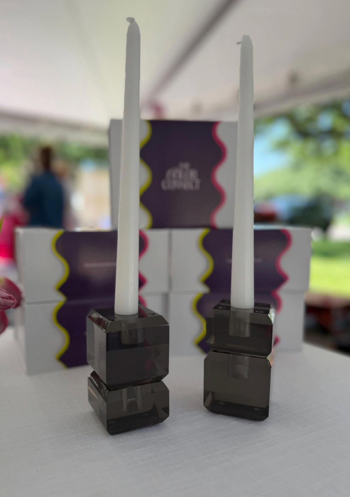 Two white taper candles in dark smoky acrylic holders, displayed in front of branded product boxes on a white table under a tent. Graphite