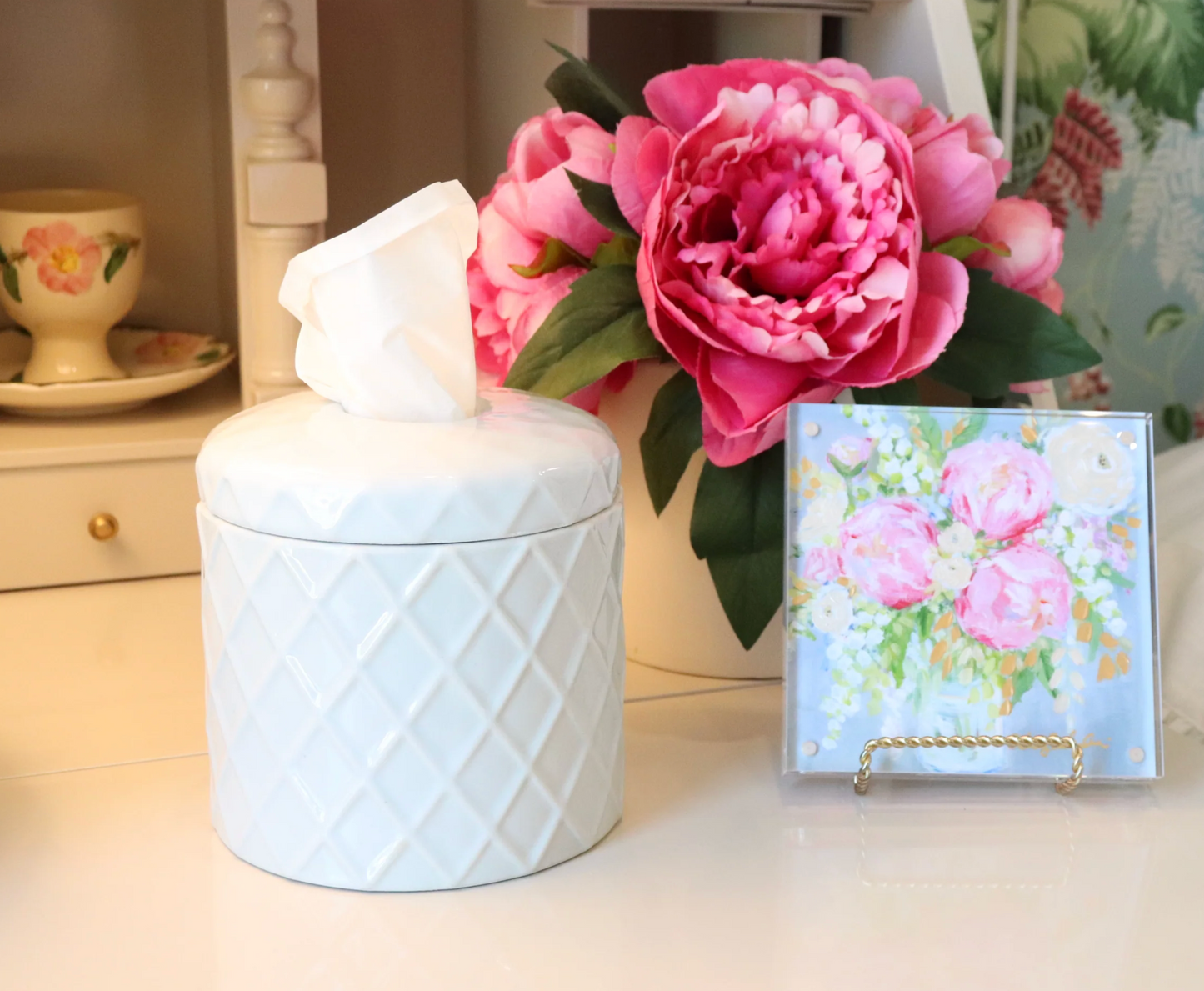 A white ceramic tissue cover with a diamond-patterned texture sits on a tabletop next to a small floral card and a pink peony flower arrangement.