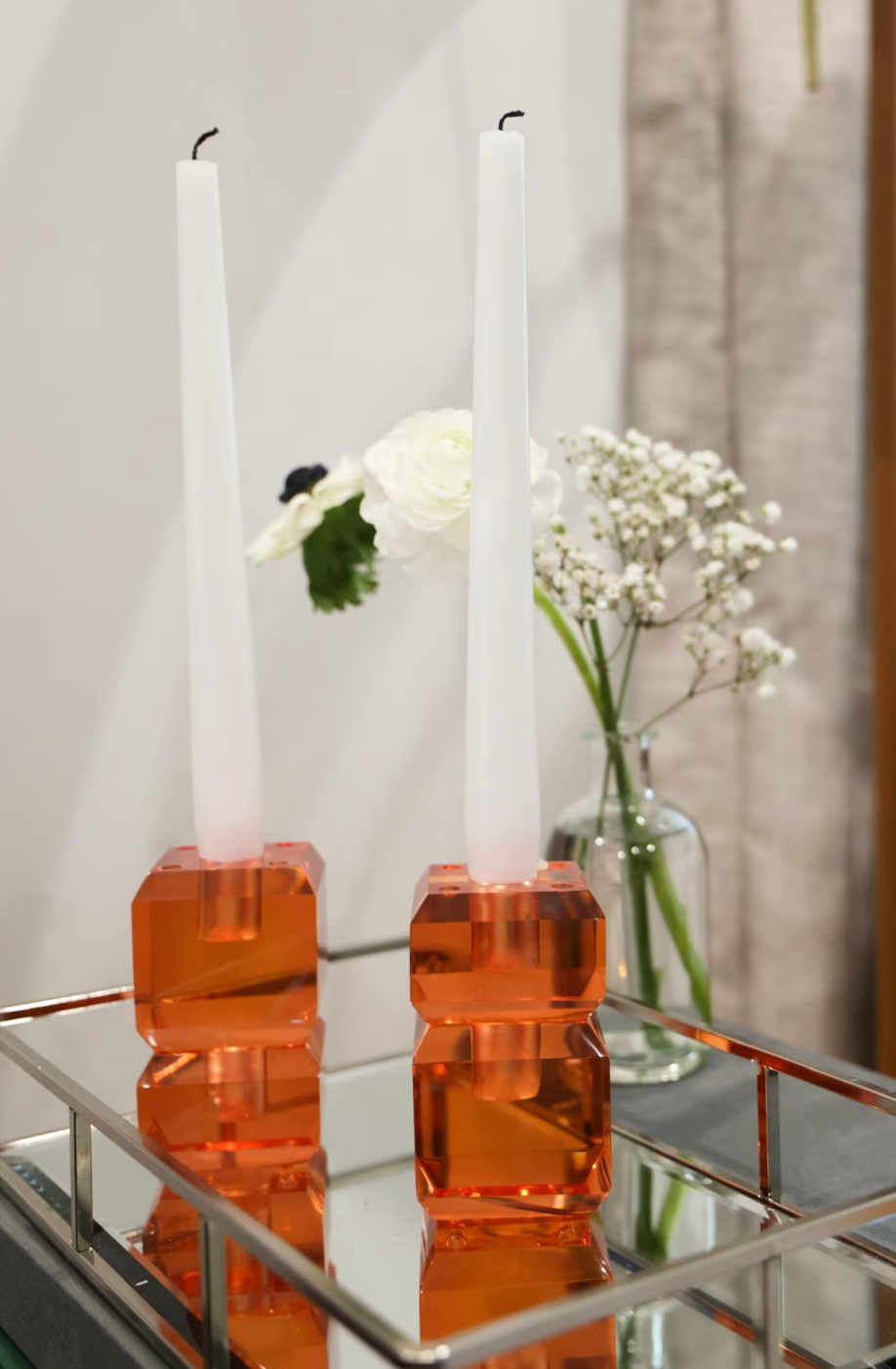 Two tall white taper candles in faceted amber-orange glass holders, displayed on a mirrored tray with white flowers in the background.