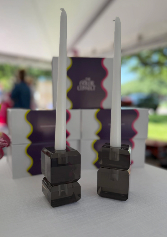 Two white taper candles in dark smoky acrylic holders, displayed in front of branded product boxes on a white table under a tent. Graphite