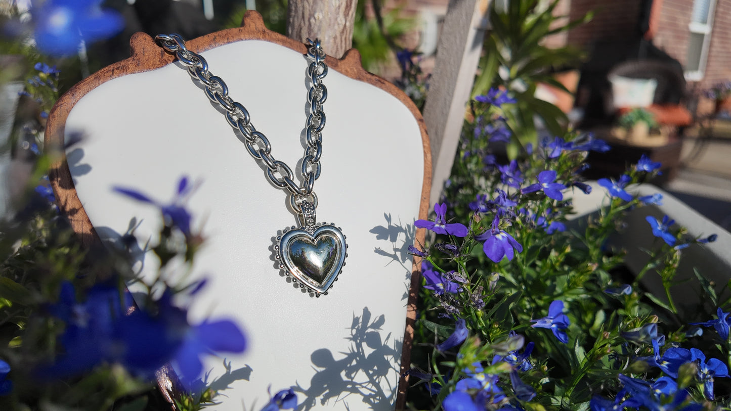 Heart-shaped silver necklace on a wooden stand with blue flowers in the background
