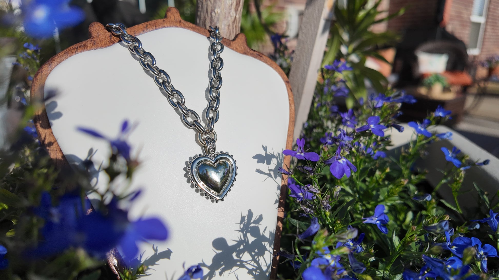 Heart-shaped silver necklace on a wooden stand with blue flowers in the background