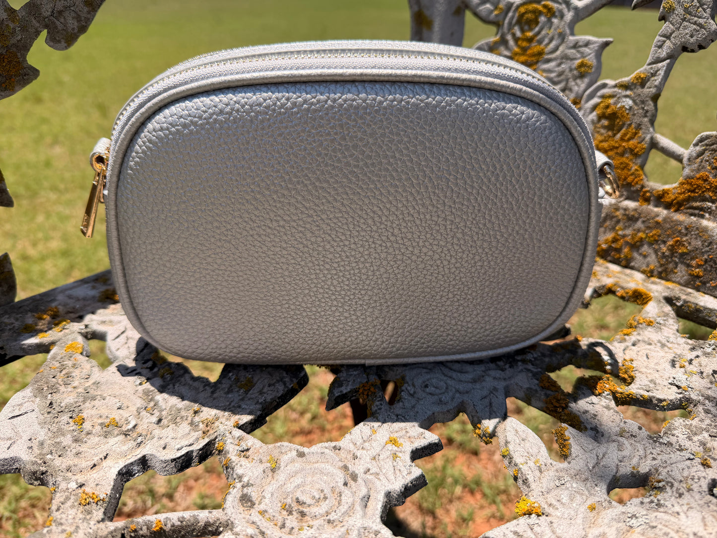 The Elodie Pebble Crossbody in Moonlit Silver styled outdoors on an ornate floral bench with gold hardware details.