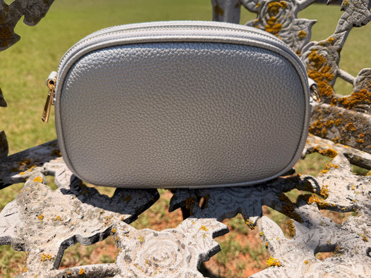 The Elodie Pebble Crossbody in Moonlit Silver styled outdoors on an ornate floral bench with gold hardware details.